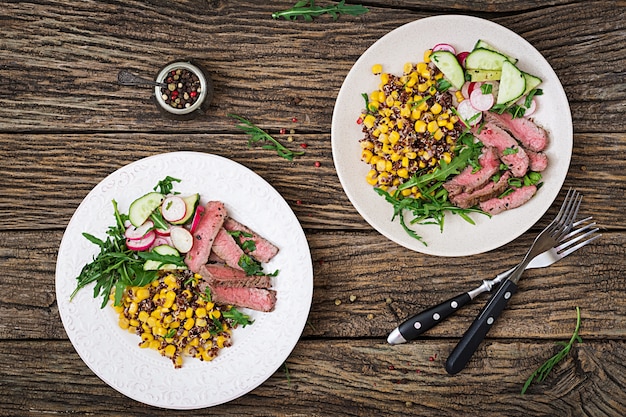 Cena Saludable. Tazón de almuerzo con filete de ternera a la parrilla y quinua, maíz, pepino, rábano y rúcula en mesa de madera. Ensalada de carne Endecha plana. Vista superior