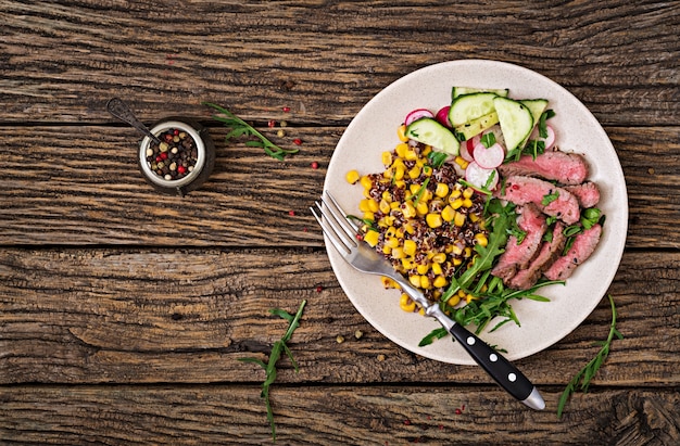 Cena Saludable. Tazón de almuerzo con filete de ternera a la parrilla y quinua, maíz, pepino, rábano y rúcula en mesa de madera. Ensalada de carne Endecha plana. Vista superior