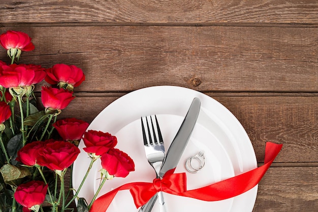 Cenário de mesa de jantar do dia dos namorados com fita vermelha, rosas, faca e garfo e anel sobre fundo de carvalho. Natureza morta