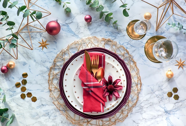 Cenário de mesa de Natal com guardanapo vermelho, poinsétia, utensílios de ouro e folhas de eucalipto em fundo de mármore. Postura plana na mesa com talheres de ouro, pratos elegantes e hexágonos geométricos.