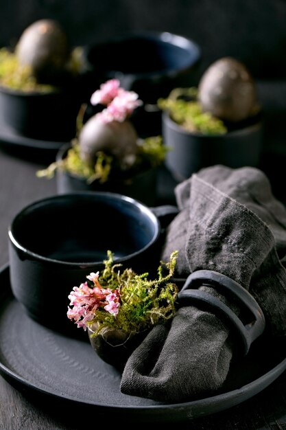 Cenário de mesa de páscoa com pratos vazios de cerâmica preta e canecas decoradas por musgo da floresta ovos de páscoa de cor preta e flores de primavera rosa com casca na mesa de madeira Feriados elegantes escuros Fechar