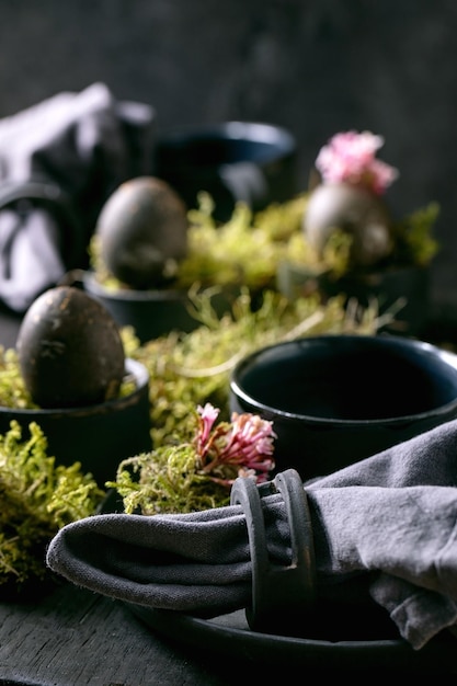 Cenário de mesa de páscoa com pratos vazios de cerâmica preta e canecas decoradas por musgo da floresta ovos de páscoa de cor preta e flores rosa da primavera na mesa de madeira Feriados elegantes escuros Fechar