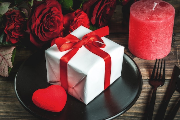 Cenário de mesa para dia dos namorados. Buquê de rosas vermelhas, amarre com uma fita vermelha, caixa de presente, corações vermelhos, vela, prato, garfo, colher e faca. Em uma mesa de madeira