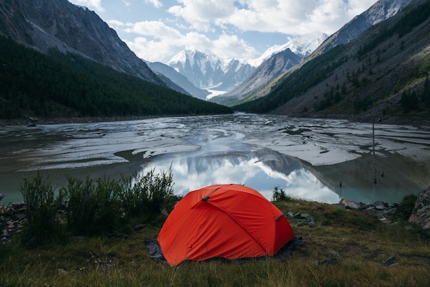 Cênica paisagem alpina com tenda laranja perto do lago de montanha lindo espelho com riachos no vale das montanhas nevadas sob céu nublado. Cenário ensolarado de alta montanha com lago glacial espelhado