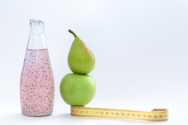 Un centímetro, una manzana, una pera y botellas de vidrio con semilla de albahaca rosa sobre un fondo blanco