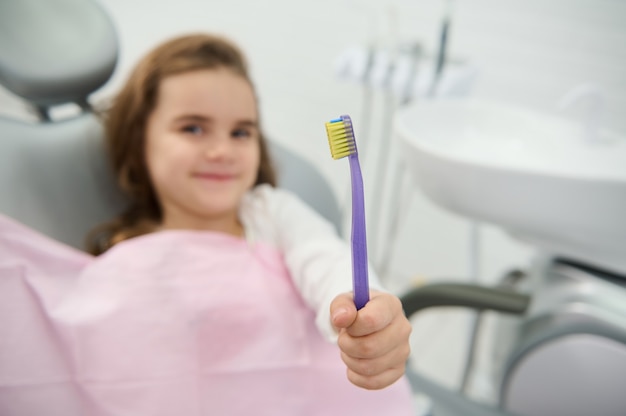 Centrarse en un cepillo de dientes en la mano de la adorable niña hermosa sonriendo tiernamente mirando a la cámara sentado en la silla del dentista durante el chequeo dental. Cuidado e higiene bucal. Concepto de odontología pediatra