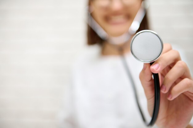 Centrarse en el fonendoscopio estetoscopio en la mano de una doctora borrosa sonriente sonrisa con dientes contra el fondo de la pared blanca con espacio publicitario de copia. Concepto de inspección médica.