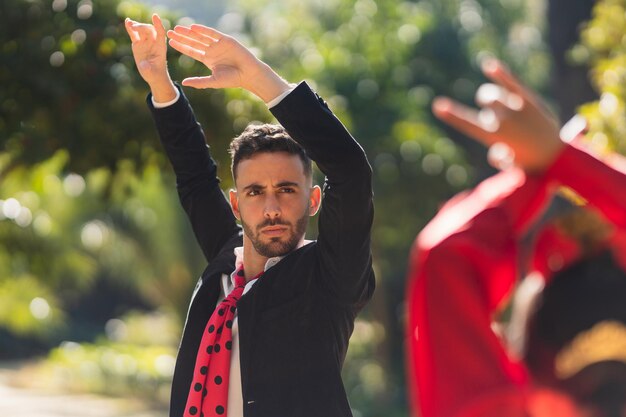 Centrarse en un hombre que levanta los brazos mientras baila flamenco con una mujer en traje de flamenca en un parque