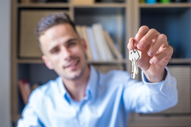 Centrarse en el manojo de llaves del apartamento de la casa en la mano del hombre sonriente. Retrato borroso del agente inmobiliario profesional del hombre confiado que ofrece la nueva unidad inmobiliaria de la vivienda al comprador potencial. De cerca