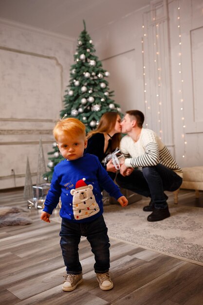 Centrarse en el niño En el fondo detrás de él, mamá y papá se están besando cerca del árbol de Navidad
