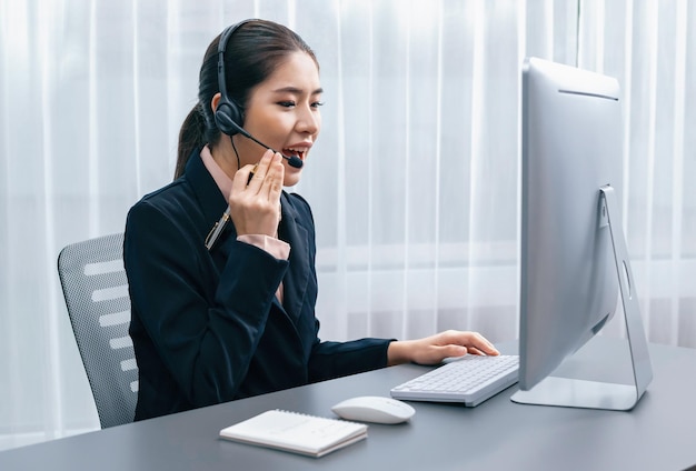 Centro de llamadas asiático entusiasta con auriculares y micrófono trabajando en su computadora portátil