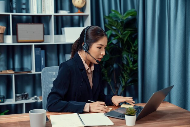 Centro de llamadas asiático entusiasta con auriculares y micrófono trabajando en su computadora portátil