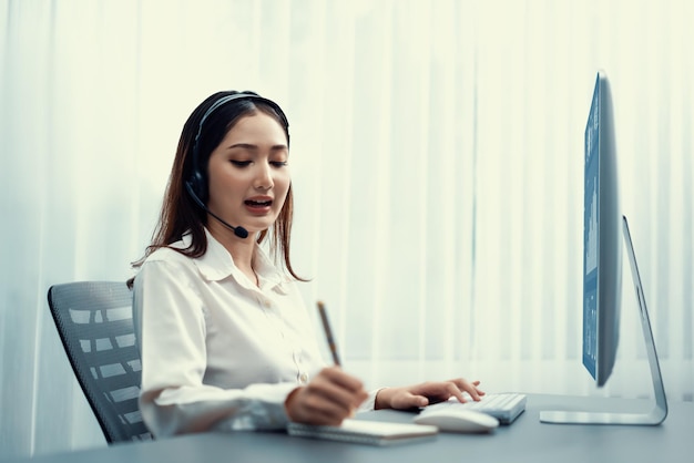 Centro de llamadas asiático entusiasta con auriculares y micrófono trabajando en su computadora portátil