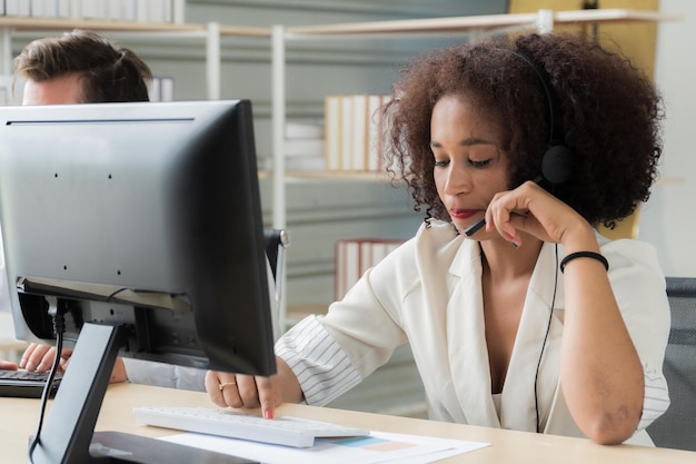 Centro de llamadas de mujer afroamericana con ropa de trabajo blanca con auriculares trabajando en el centro de llamadas en la computadora