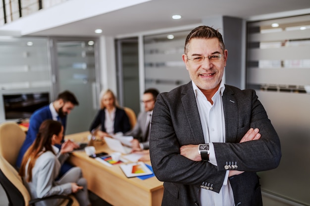 CEO caucásico acertado de mediana edad de pie en la sala de juntas con las manos cruzadas y mirando a la cámara. En segundo plano están sus empleados trabajando en un proyecto para un cliente importante.