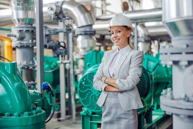 CEO femenina exitosa rubia de mediana edad en traje con casco en la cabeza de pie en la planta de energía con los brazos cruzados