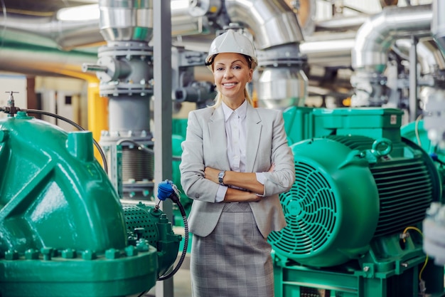 CEO im Anzug mit Helm auf dem Kopf stehend im Heizwerk mit verschränkten Armen