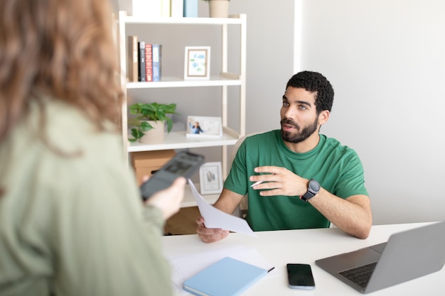 Ceo jovem e sério do Oriente Médio à mesa com laptop conversando com gerente com smartphone