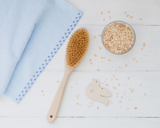 Cepillo para el cuerpo de madera, toalla azul y avena cruda sobre un fondo de madera blanca