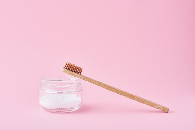 Cepillo de dientes de bambú de madera y polvo de bicarbonato de sodio en frasco de vidrio sobre un fondo rosa. Salud de los dientes y concepto de boca