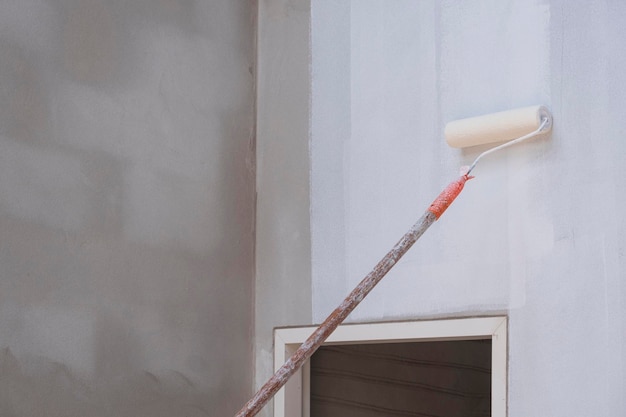 Cepillo de rodillo de mango largo que aplica pintura blanca de imprimación en la pared de cemento en el sitio de construcción de la casa