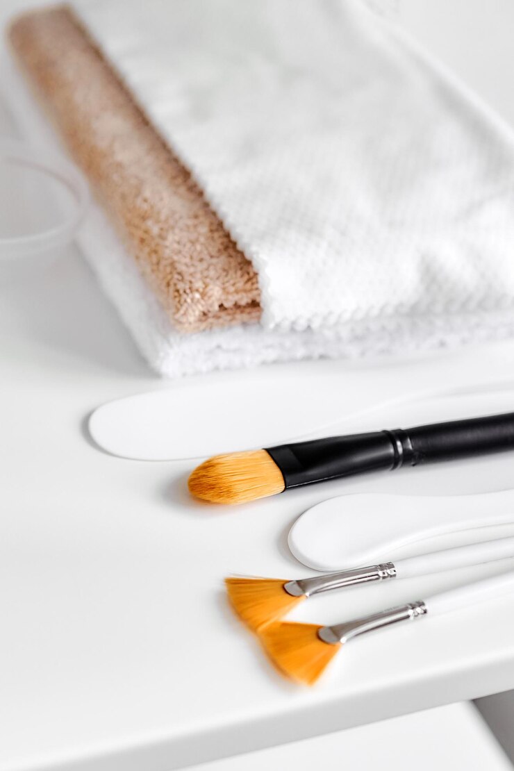 Cepillos de nailon cosméticos, espátulas de plástico y una pila de toallas dobladas en un estante blanco Cosmetología