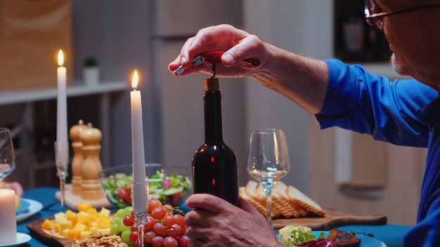 Cerca de abrir una botella de vino durante una cena romántica. Pareja de ancianos maduros sentados a la mesa en la cocina, hablando, disfrutando de la comida, celebrando su aniversario en el comedor.