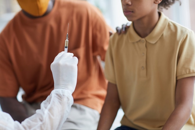 Cerca de un adolescente afroamericano mirando a la enfermera sosteniendo una jeringa con aguja durante la vacunación covid en la clínica con el padre apoyándolo