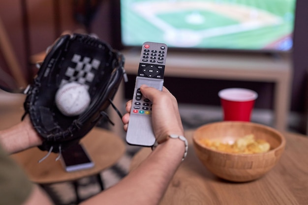 Cerca de un aficionado al béisbol sosteniendo TV revpmte mientras ve el partido en casa