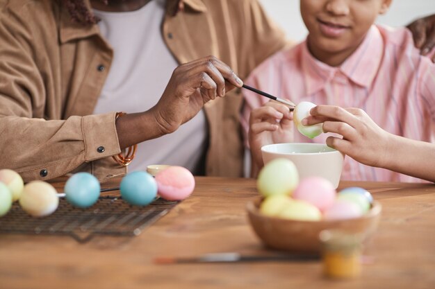 Cerca de amorosa madre e hija afroamericana pintando huevos de Pascua juntos mientras está sentado en la mesa de madera en el acogedor interior de una casa