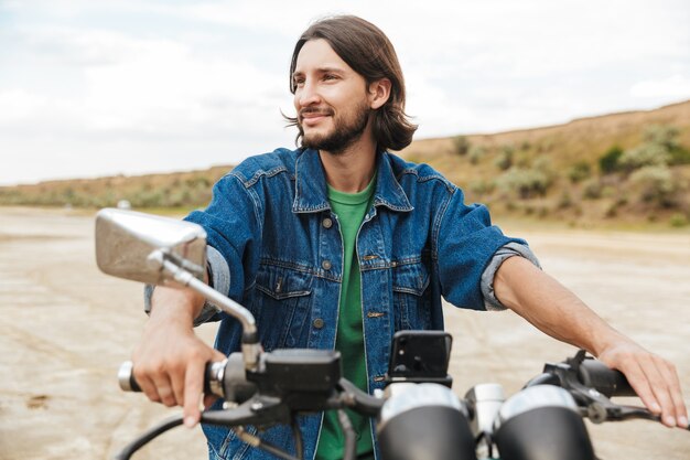 Cerca de un apuesto joven feliz vistiendo ropa casual sentado en una motocicleta en la playa