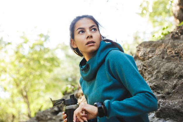 Cerca de una atractiva joven mujer fitness vistiendo ropa deportiva haciendo ejercicio al aire libre, sosteniendo una botella de agua, mirando su reloj