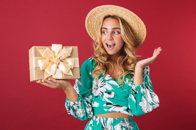 Cerca de una atractiva joven rubia feliz con vestido de verano y sombrero de paja que se encuentran aisladas sobre la pared rosa, mostrando el cuadro actual