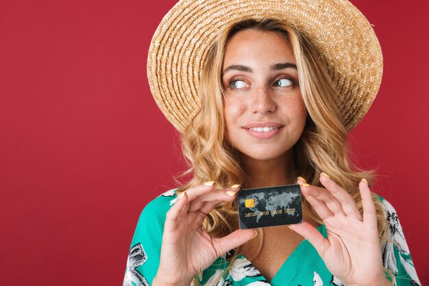Cerca de una atractiva joven rubia sonriente con vestido de verano y sombrero de paja que se encuentran aisladas sobre la pared rosa, mostrando la tarjeta de crédito