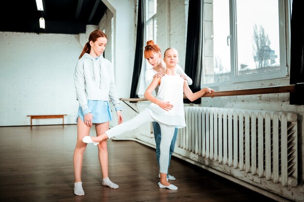 Cerca de la barra de ballet. Adolescente rubio de pelo largo con ropa deportiva blanca haciendo ejercicio cerca de la barra de ballet