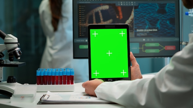 Cerca del bioquímico sentado en el lugar de trabajo en el laboratorio usando una tableta de pantalla de maqueta verde con pantalla chroma key. compañero de trabajo que trabaja en segundo plano trayendo muestra de sangre.