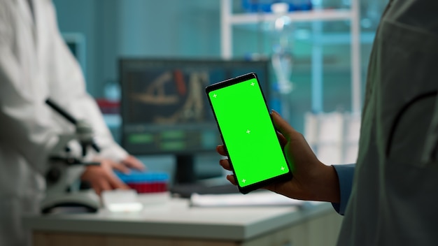 Cerca del bioquímico sosteniendo y mirando el teléfono con pantalla chroma key de pie en el laboratorio biológico mientras la enfermera en bata blanca trae una muestra de sangre. Científico con teléfono con maqueta, pantalla verde