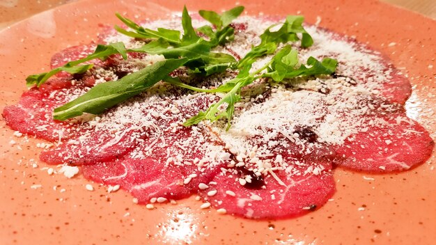 Cerca de carpaccio de carne con parmesano y rúcula en un plato grande de cerámica naranja sobre una mesa de madera. Con aceite de oliva.