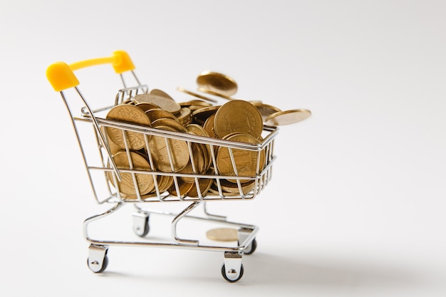 Cerca del carrito de supermercado para compras con mango de plástico amarillo con monedas de oro cayendo en él aislado sobre fondo blanco. Concepto de compras. Copie el espacio para publicidad.