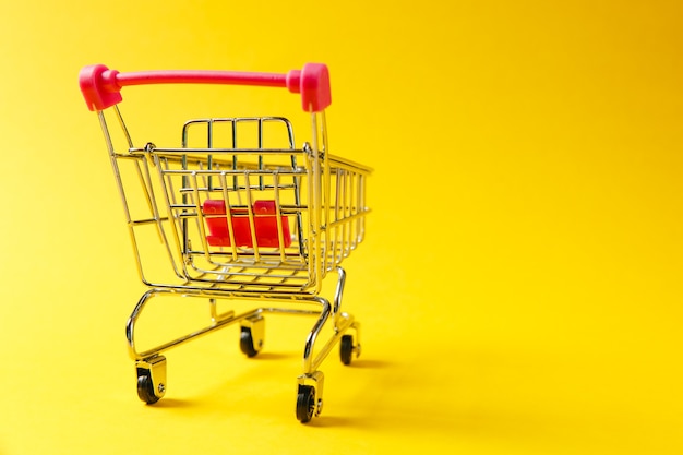 Cerca del carrito de supermercado para ir de compras con ruedas negras y elementos de plástico rojo en el mango aislado sobre fondo amarillo. Concepto de compras. Copie el espacio para publicidad.