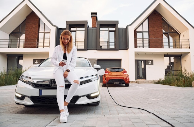 Cerca de la casa, una mujer joven vestida de blanco está con su auto eléctrico durante el día.