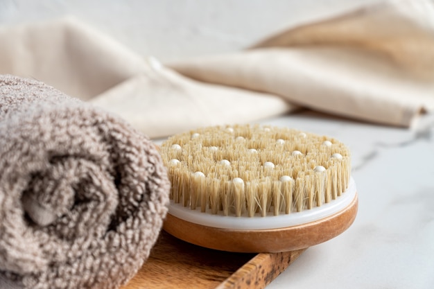 Cerca de un cepillo de bambú de baño para masaje en seco y una toalla de algodón en una bandeja de madera sobre mármol