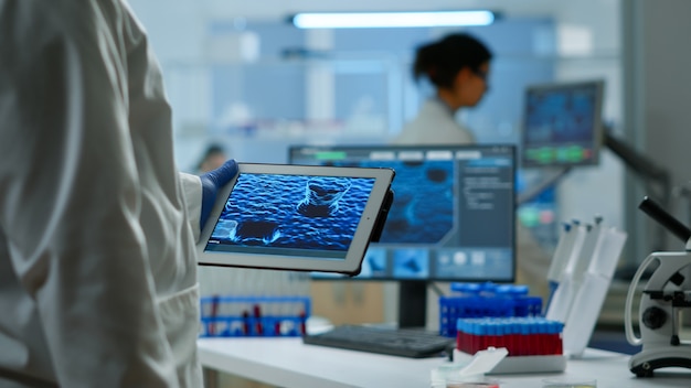 Cerca de científico en bata de laboratorio trabajando sosteniendo tableta de pie en el moderno laboratorio equipado. Químicos del equipo que examinan la evolución de las vacunas utilizando herramientas de alta tecnología y química para el desarrollo de virus