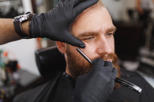 Cerca del cliente de servicio de peluquería profesional masculina, afeitado de barba grande y gruesa con navaja recta