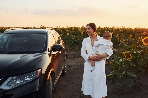 Cerca de un coche negro moderno La joven madre con su pequeño hijo está al aire libre en el campo agrícola Hermoso sol