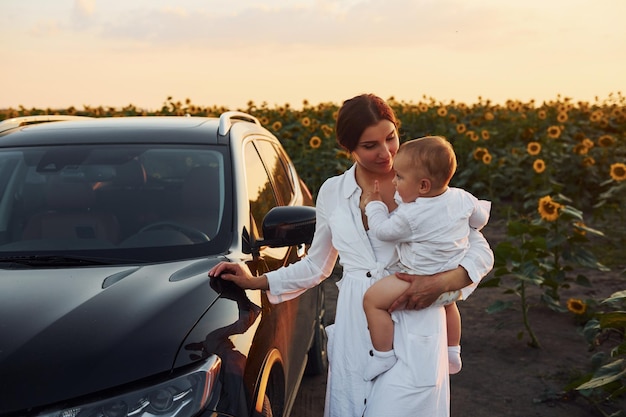 Cerca de un coche negro moderno La joven madre con su pequeño hijo está al aire libre en el campo agrícola Hermoso sol