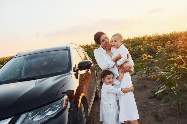 Cerca de un coche negro moderno La joven madre con su pequeño hijo y su hija están al aire libre en el campo agrícola Hermoso sol