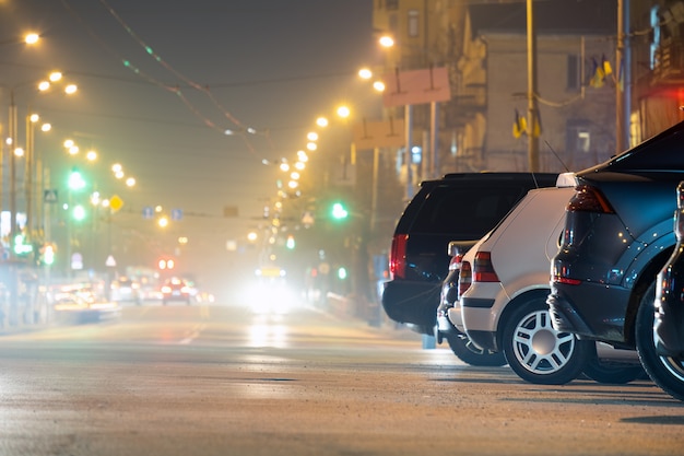 Cerca de los coches aparcados en la carretera por la noche con vista borrosa de los semáforos de vehículos en movimiento en las calles de la ciudad.