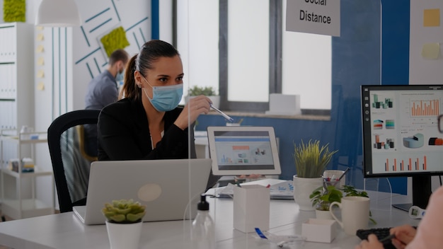 Cerca de compañeros de trabajo con mascarilla trabajando juntos en un proyecto financiero usando una tableta mientras están sentados en la oficina de la empresa. El equipo mantiene la distancia social para evitar la infección con covid19