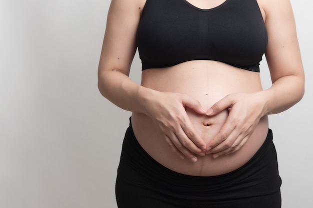 Cerca del cuerpo central de la mujer embarazada con sujetador negro muestra su mano simboliza el corazón en su estómago, retrato de luz de estudio y espacio de copia en blackground blanco